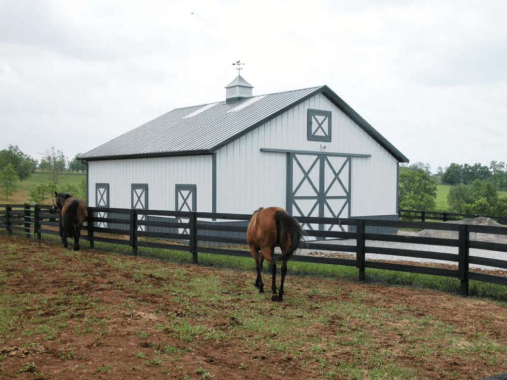 Post-frame horse barn with crossbuck sliding doors and black fence in front.