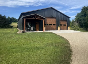 Shop with timber frame porch featuring green steel siding, exposed wood trusses, and dual overhead garage doors in Wisconsin