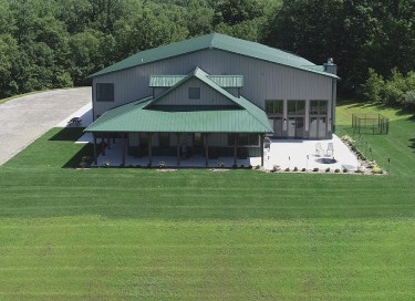 Luxury hunting cabin with green steel roof, covered porch, and large equipment storage building in wooded setting