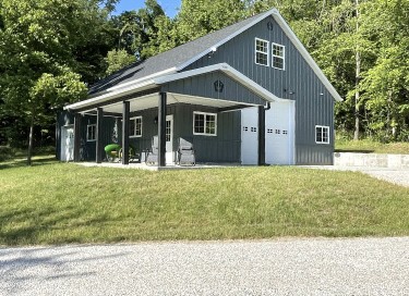 Garage with living quarters featuring gray steel siding, covered porch, overhead garage door, and loft space in rural settingQuarters