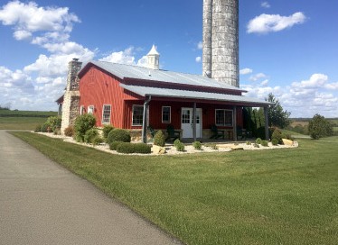 Pole barn house with silo featuring red steel siding, covered porch, stone chimney, and rural setting in Kentucky