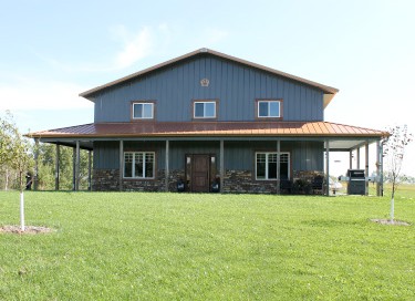 Pole barn house with blue steel siding, covered wraparound porch, and stone wainscot in Wisconsin