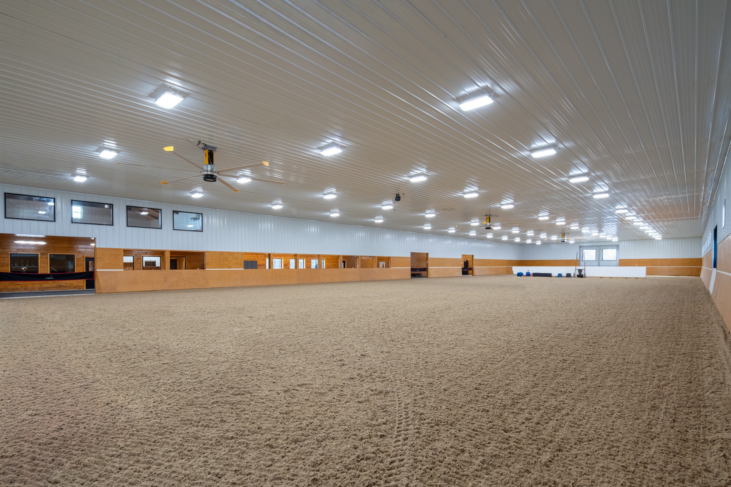 Indoor riding arena at The Settlement Equestrian Center with bright overhead lighting, ceiling fans, and groomed equine footing