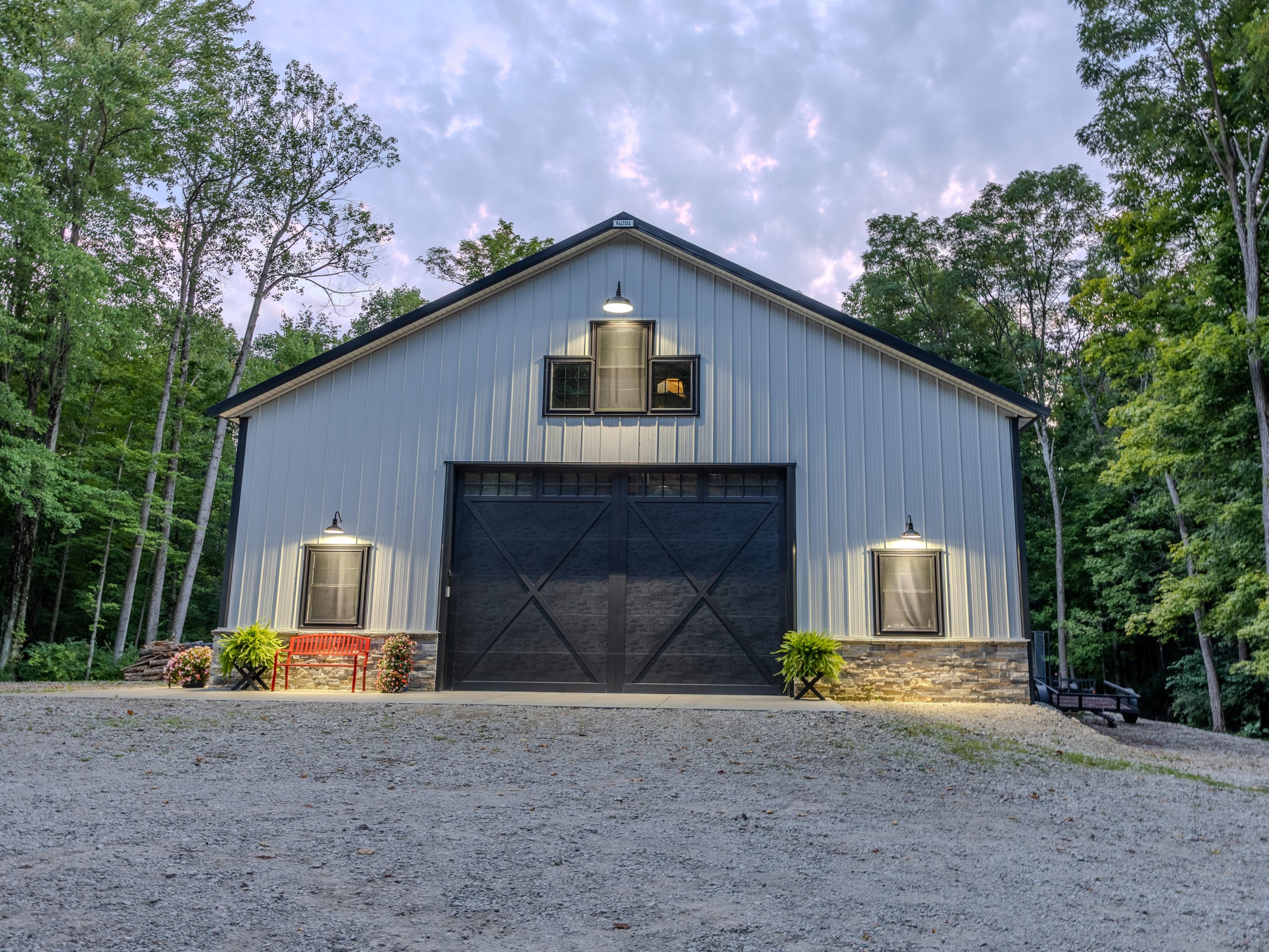 Dusk view of custom pole barn with illuminated gooseneck lights, black garage door and stone wainscot