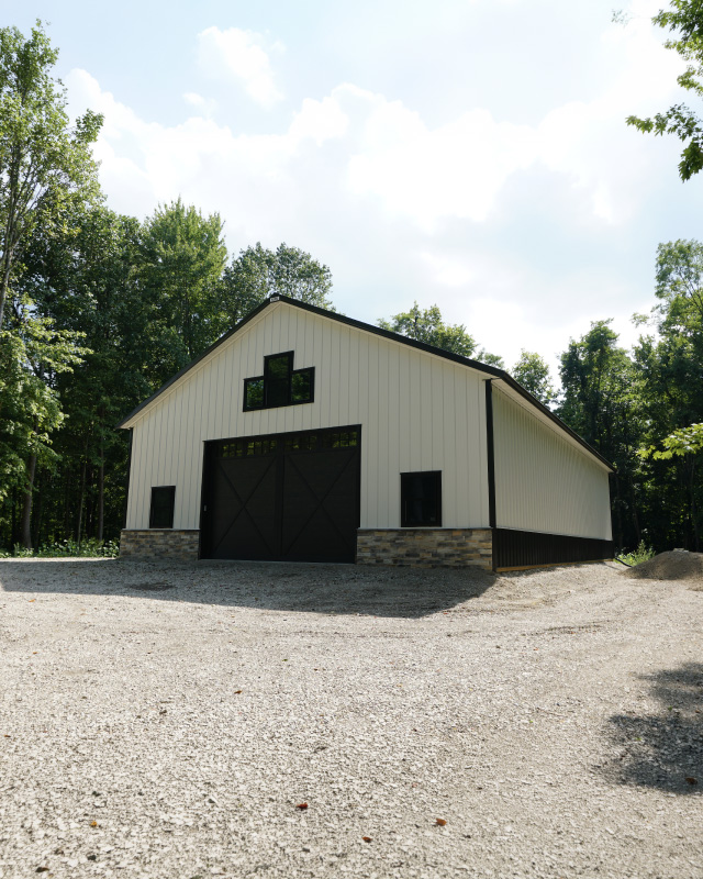 White post-frame storage building with black trim, cupola, and metal roof on gravel lot in wooded setting