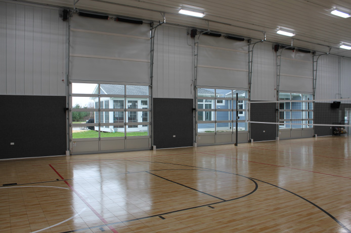 Interior basketball court with hardwood flooring, high ceilings, and overhead doors inside post-frame building Interior basketball court with hardwood flooring, high ceilings, and overhead doors inside post-frame building