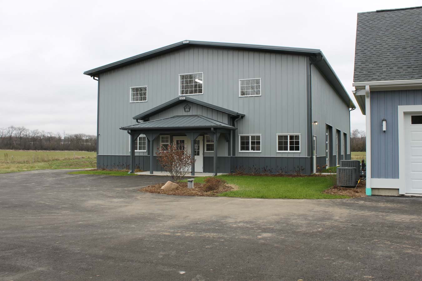 Project #94-0512 Exterior of indoor basketball court building with gray metal siding, covered entry, and residential driveway in Wisconsin