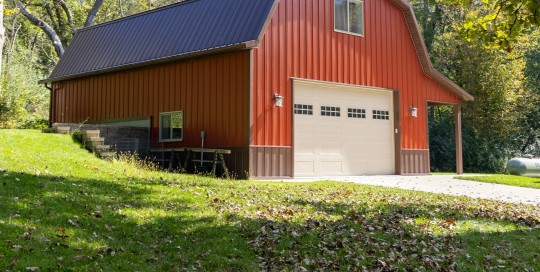 Two-story gambrel pole barn garage with red steel siding and brown trim on a wooded hillside in Nerstrand, Minnesota