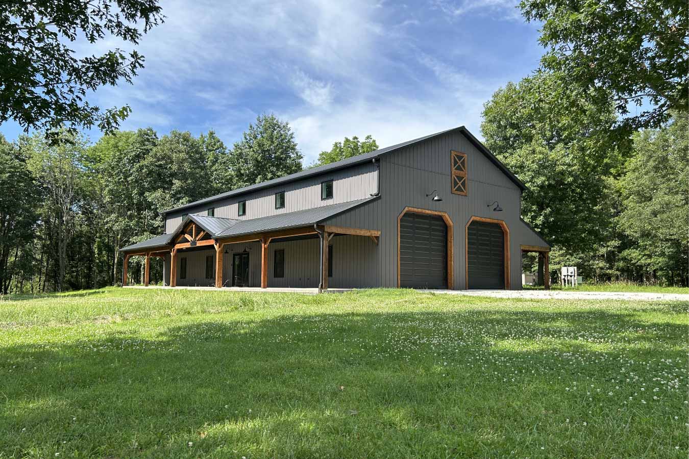 Front exterior of a custom post-frame barndominium featuring dark gray vertical siding, two large overhead garage doors with cedar-stained trim, bronze roof, and a covered front porch supported by cedar-wrapped posts. Surrounded by green lawn and trees under a blue sky. Built by Cornerstone Construction in partnership with Walters Buildings.