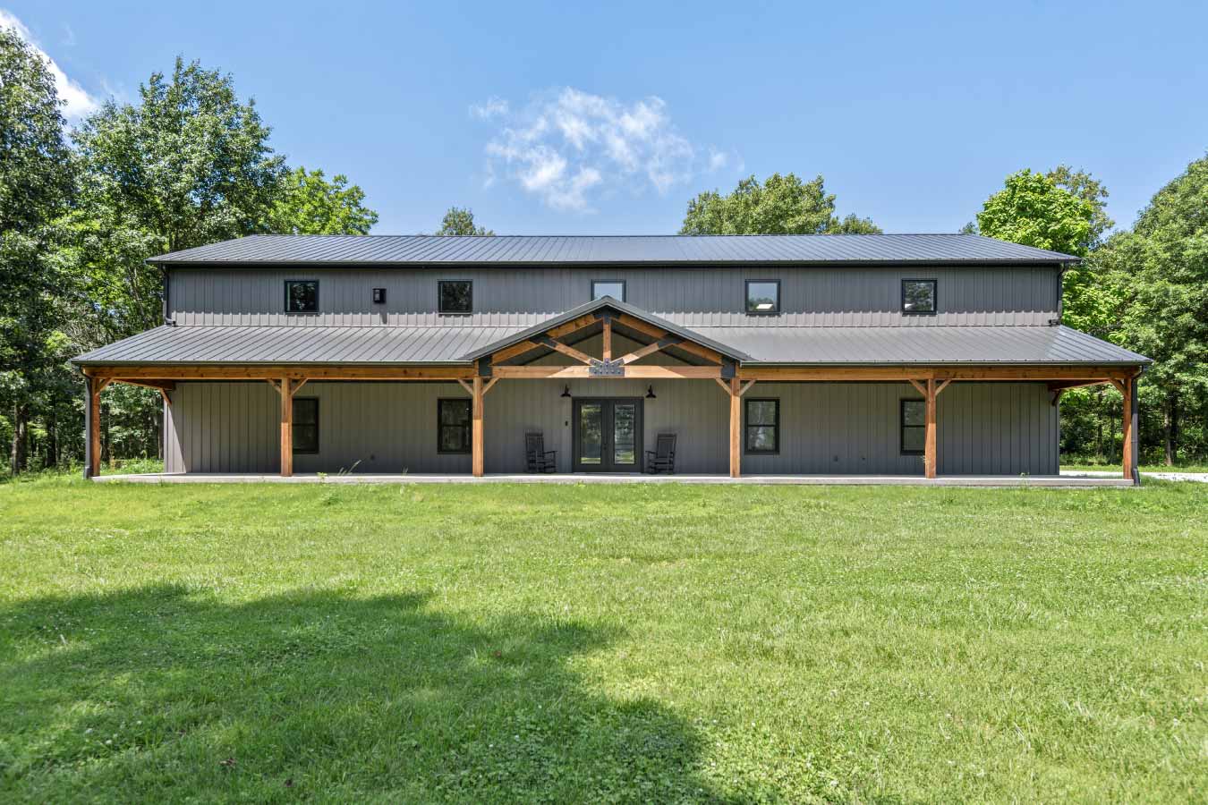 Modern two-story barndominium with full-length covered porch, cedar-wrapped posts, and dark metal roof, set on a private wooded lot.