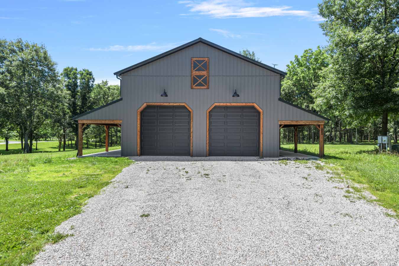 Front view of a modern barndominium garage with two large black overhead doors framed in cedar trim, flanked by covered side porches. The structure features a dark metal roof and vertical steel siding, set on a gravel driveway and surrounded by mature trees.