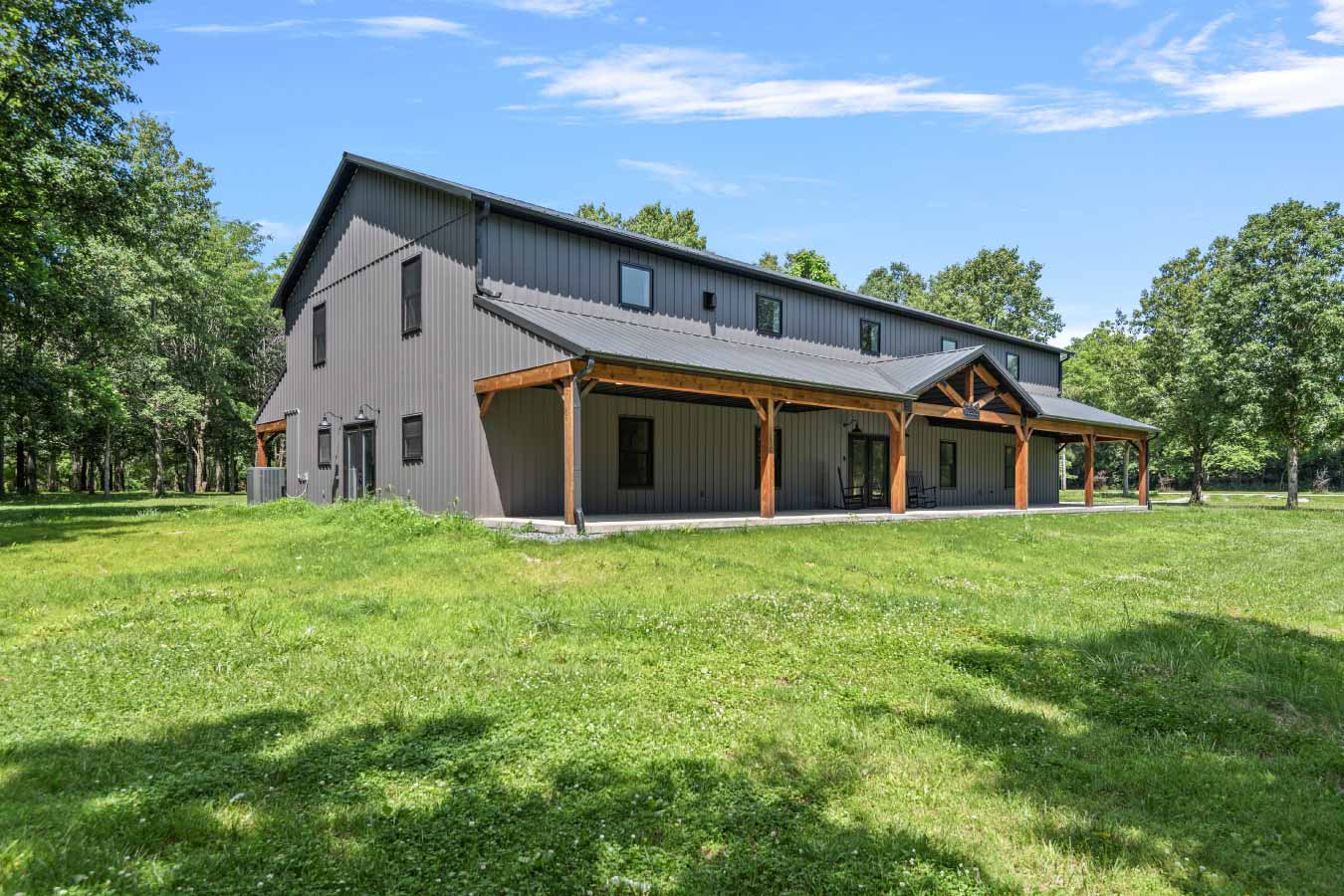 Angled view of a modern post-frame barndominium with a dark metal roof, vertical steel siding, and full-length covered front porch with cedar-wrapped posts. Surrounded by open lawn and mature trees on a private 2.48-acre lot in Illinois, this two-story home combines rural charm with contemporary design. Built by Cornerstone Construction Services.