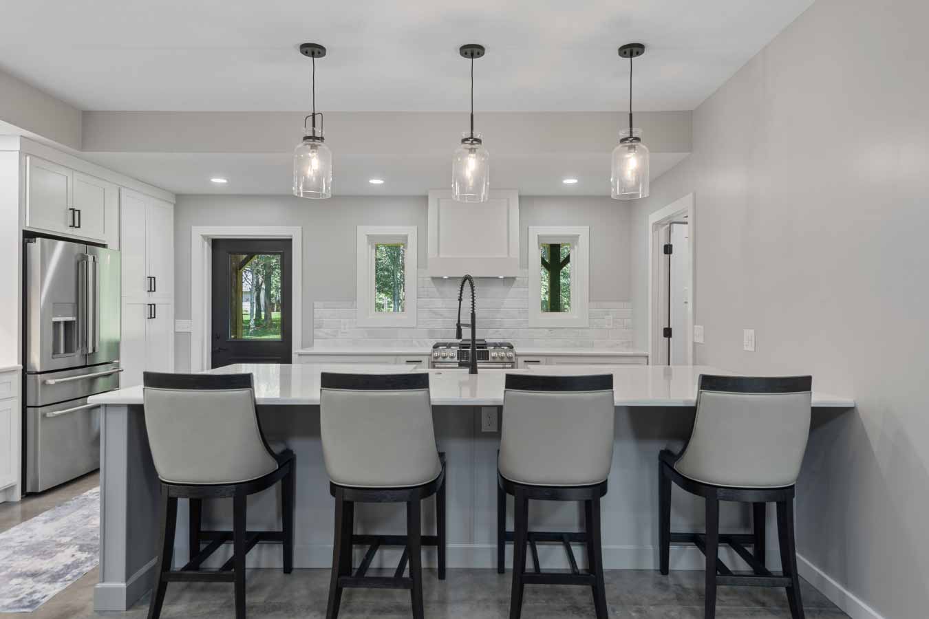 Front-facing view of a modern kitchen island with seating for five, quartz countertops, and pendant glass lighting. The island features a deep farmhouse sink with a matte black industrial-style faucet, overlooking a stainless steel gas range and a white tile backsplash. Large windows and a rear entry door provide views of the wooded backyard in this custom-built barndominium by Cornerstone Construction Services.