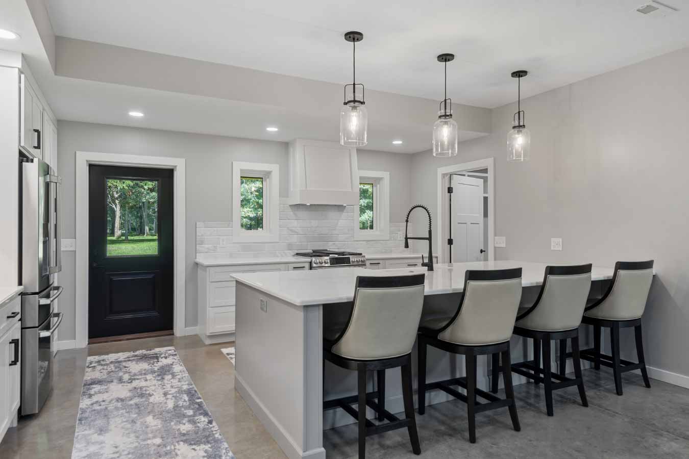 Wide-angle view of a modern kitchen with a large island and bar seating for five. Features include a quartz countertop, pendant glass lights, a black gooseneck faucet, gas range with white hood, stainless steel refrigerator, and a black back door with views of a wooded yard.