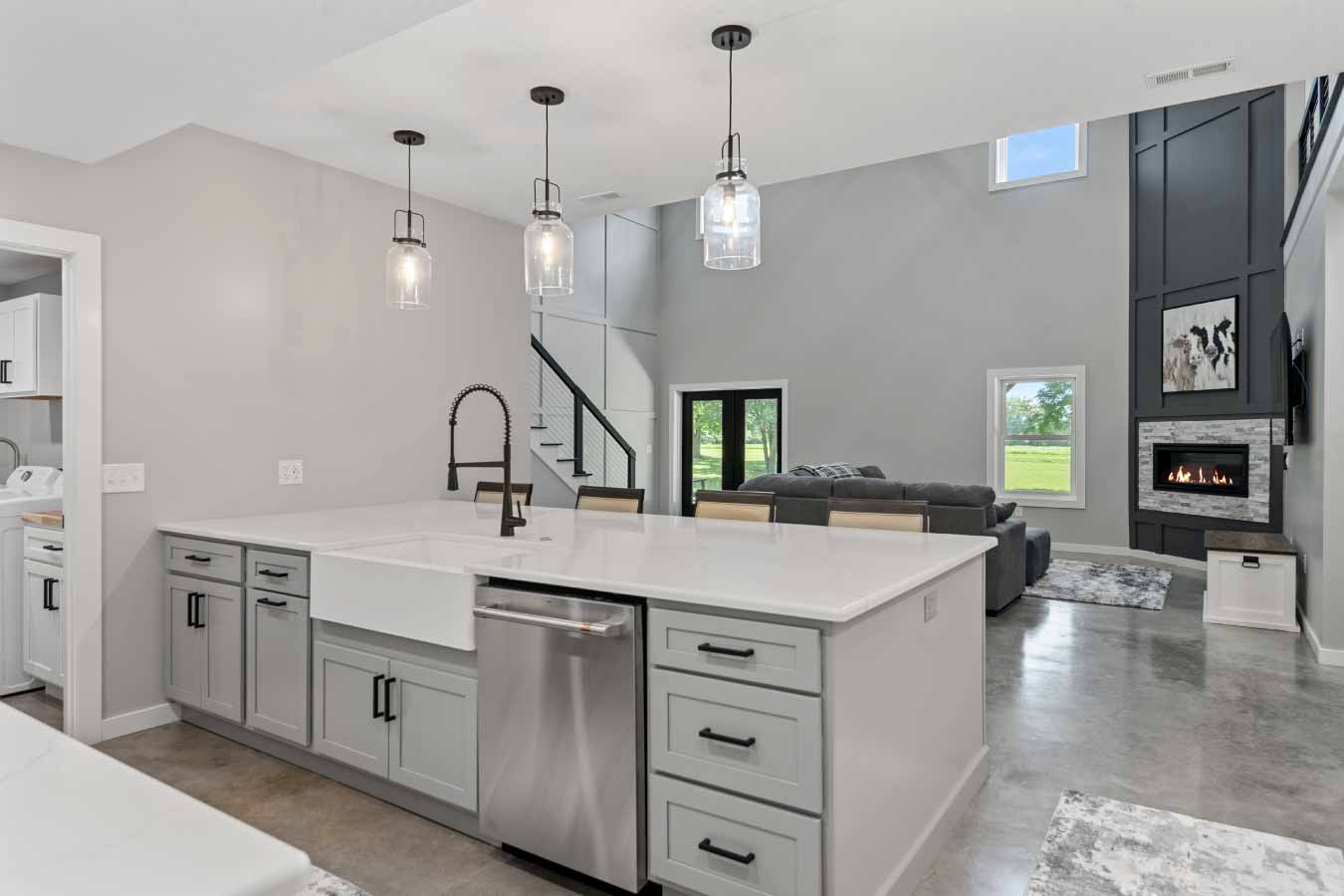 View from the kitchen island looking into the open-concept great room of a modern barndominium. Features include a large quartz island with farmhouse sink and matte black faucet, polished concrete floors, and a soaring two-story living room with a tall paneled fireplace, built-in gas insert, and large windows overlooking a private wooded lot.