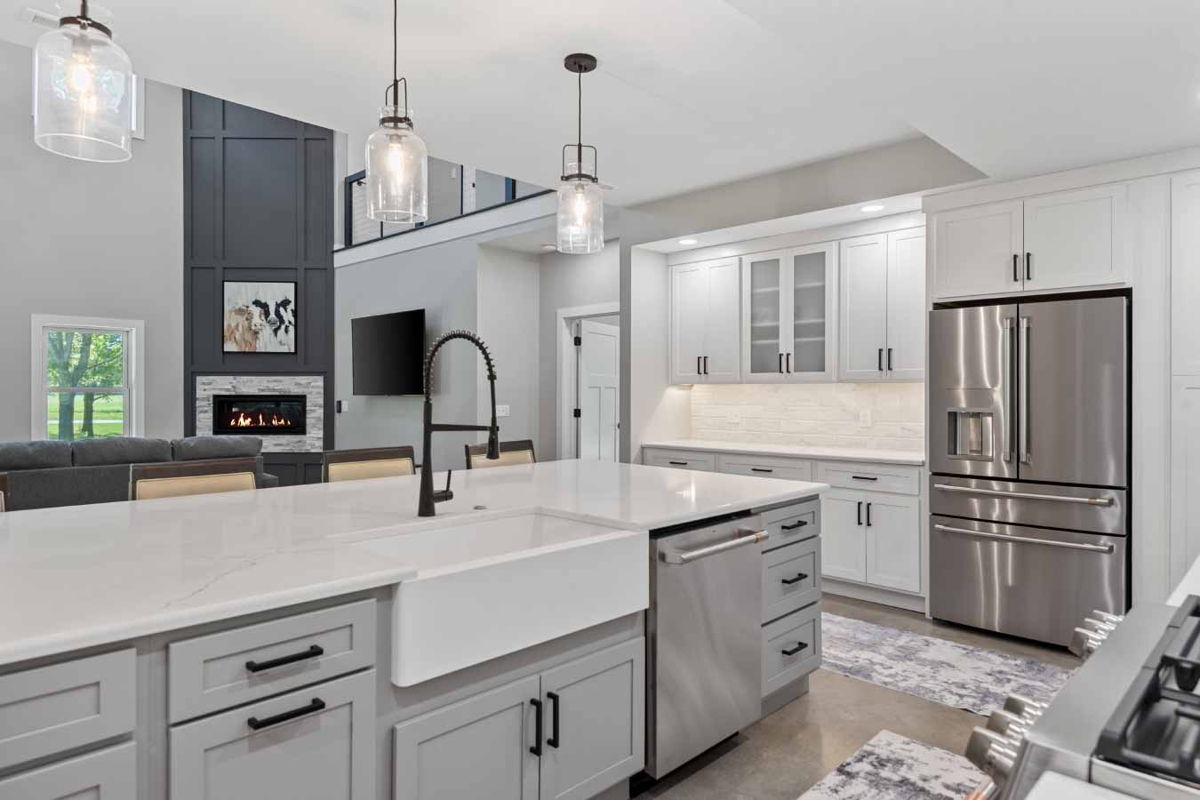 View from the kitchen island featuring a white farmhouse sink and matte black faucet, looking toward the master bedroom entry. The kitchen includes quartz countertops, gray shaker cabinets, stainless steel appliances, and glass pendant lighting. A two-story great room with a floor-to-ceiling fireplace and polished concrete floors adds to the upscale aesthetic of this post-frame barndominium.