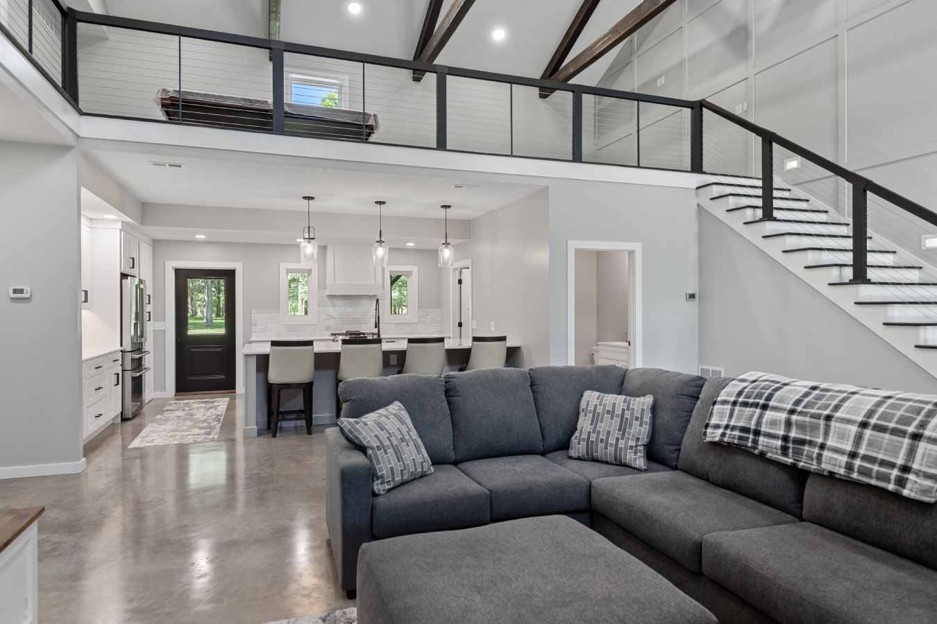 Spacious great room in a modern barndominium with polished concrete floors, vaulted ceilings, exposed wood beams, and a lofted second floor with metal cable railing. The view includes a large sectional sofa, staircase with integrated lighting, and a clear sightline into the kitchen and island seating area.