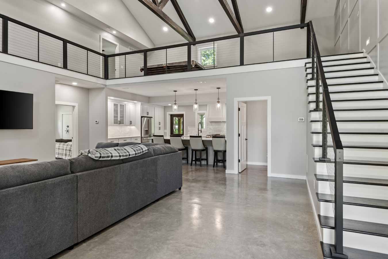 Wide interior view of a modern barndominium showcasing the open-concept great room, large sectional sofa, exposed beam ceiling, cable railing loft, and polished concrete floors. The image includes the open staircase with white risers and dark treads, a view into the half bathroom, and the adjacent kitchen with bar seating and pendant lighting.