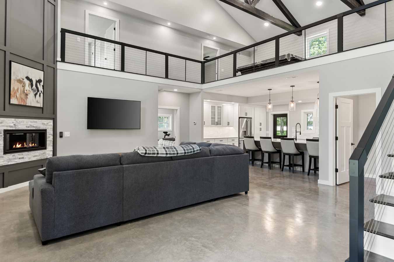 Wide-angle interior view of a luxury barndominium featuring an open-concept great room with polished concrete floors, vaulted ceilings with exposed beams, and a loft with cable railing. The layout includes a large sectional sofa facing a floor-to-ceiling paneled fireplace with a built-in gas insert, adjacent to a modern kitchen with a large island, pendant lighting, and bar seating. Also visible is a staircase with black railings and white risers.