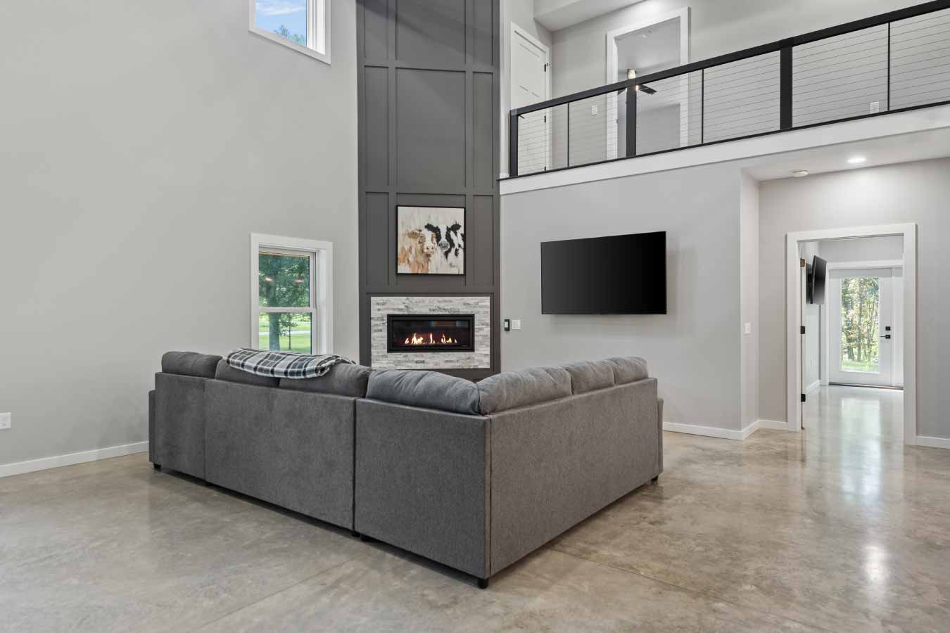 Living room of a modern barndominium featuring a built-in gas fireplace with stacked stone surround, tall paneled accent wall, and mounted television. A large gray sectional anchors the space, with polished concrete floors and a double-height ceiling leading to an upper loft with cable railing. A doorway opens to a secondary living space with natural light and views of the wooded property.