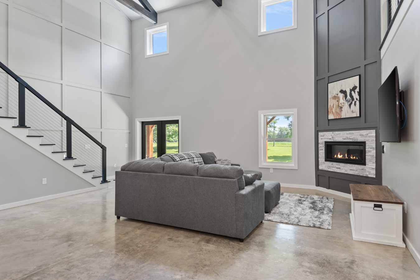 Open-concept great room with polished concrete floors, a large gray sectional, and a modern gas fireplace. A glass door under the covered front porch provides an exterior entrance, while multiple windows and high ceilings flood the space with natural light. A black cable-railed staircase and decorative wall paneling add to the modern farmhouse aesthetic.