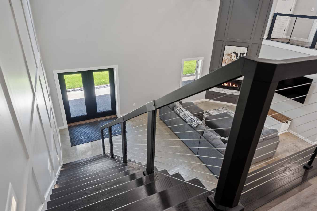 Wide-angle interior view of a luxury barndominium featuring an open-concept great room with polished concrete floors, vaulted ceilings with exposed beams, and a loft with cable railing. The layout includes a large sectional sofa facing a floor-to-ceiling paneled fireplace with a built-in gas insert, adjacent to a modern kitchen with a large island, pendant lighting, and bar seating. Also visible is a staircase with black railings and white risers.