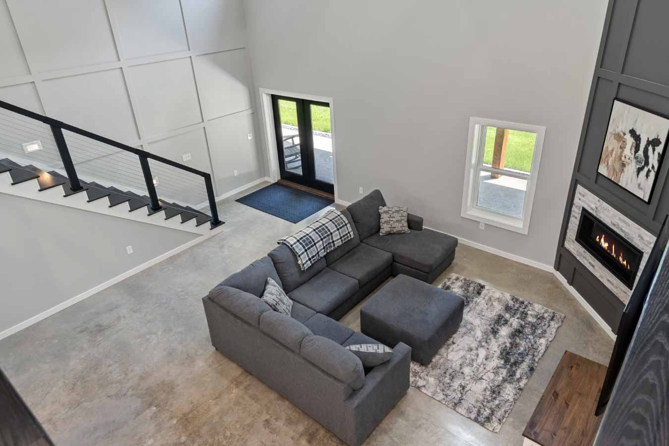Looking down from the loft onto the great room featuring a cozy gray sectional, gas fireplace with vertical trim, and double black doors leading outside. A modern staircase with black cable railing runs along the wall, and the polished concrete floor adds an industrial-modern touch.