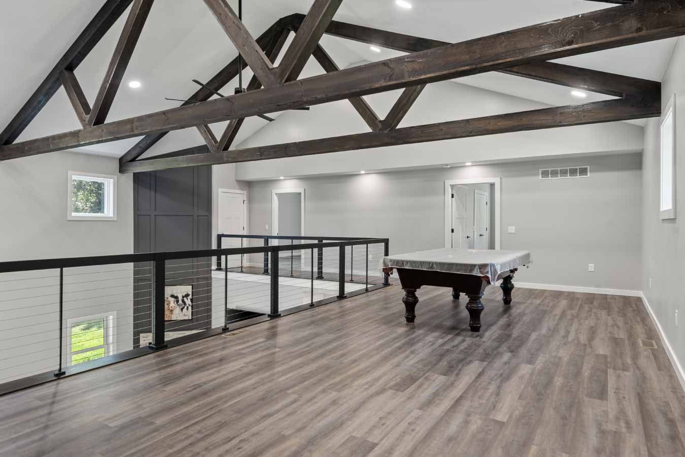 Spacious loft with vaulted ceilings, exposed timber trusses, and a pool table, overlooking the great room below. Black cable railing lines the open walkway, and two doors lead to upstairs bedrooms.