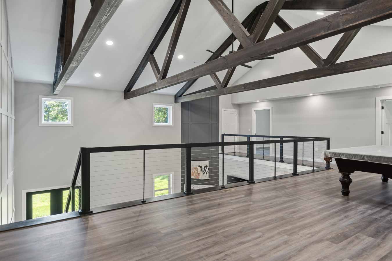 Wide view of open loft with vaulted ceilings, exposed wood trusses, and modern cable railing. Overlooks great room with large windows and entry doors below.
