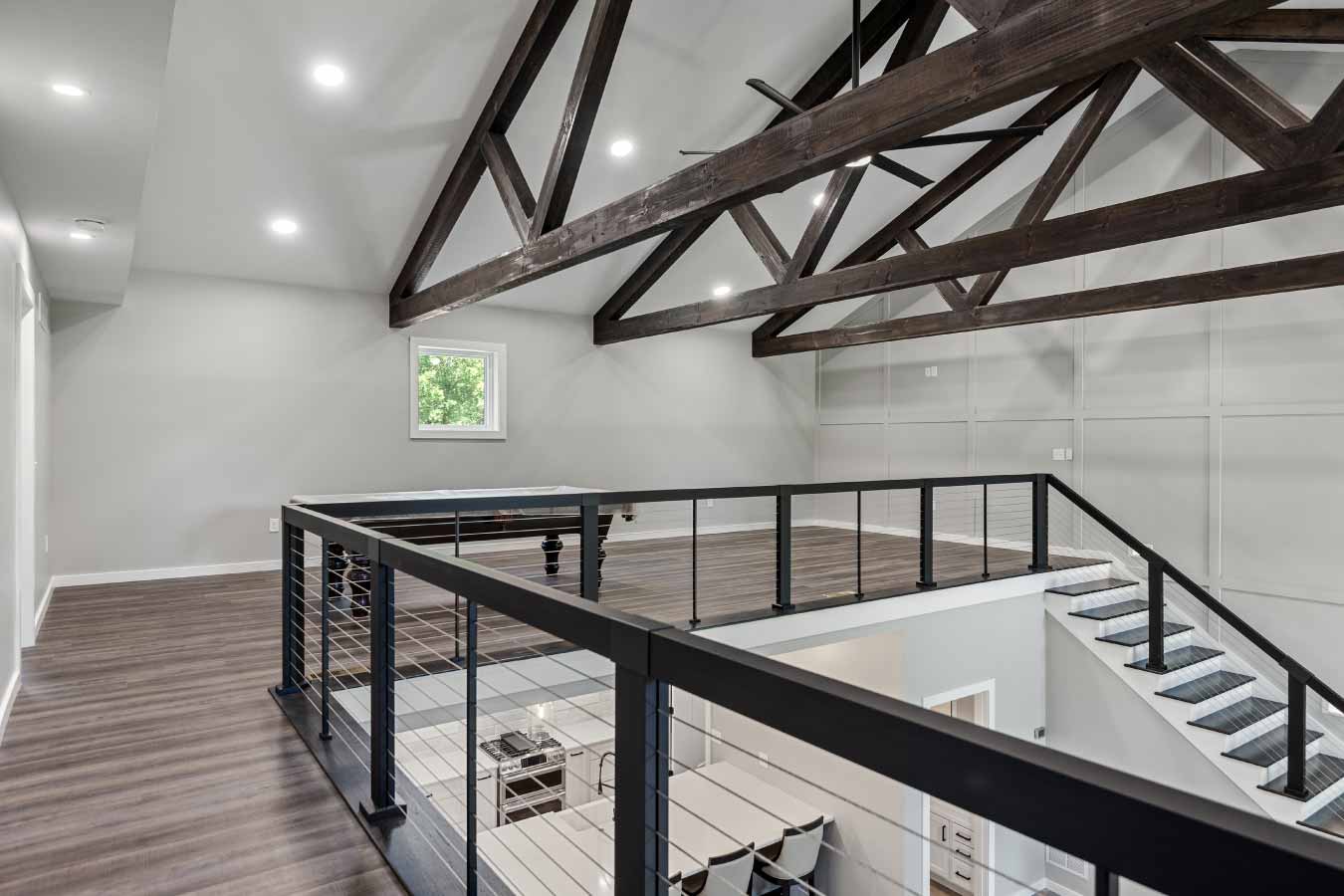 View from upstairs bedroom overlooking open loft in a modern barndominium. Features exposed wood trusses, cable railing, and a view down to the kitchen in this custom post frame home.