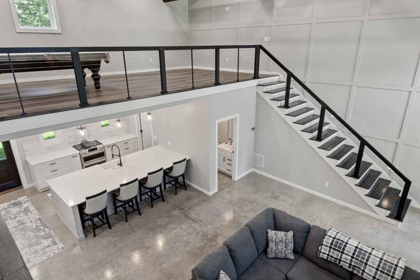 View from loft overlooking open-concept barndominium interior. Features modern cable railing, floating staircase, spacious kitchen island with seating, and polished concrete floors in this custom post frame home.