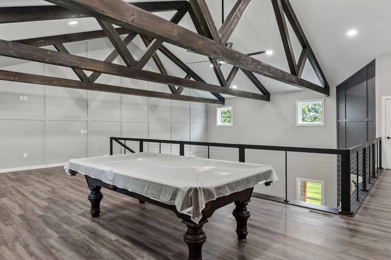 Spacious loft recreation area in a modern barndominium featuring a covered pool table, exposed wood trusses, cable railing, and luxury vinyl plank flooring in this custom post frame home.