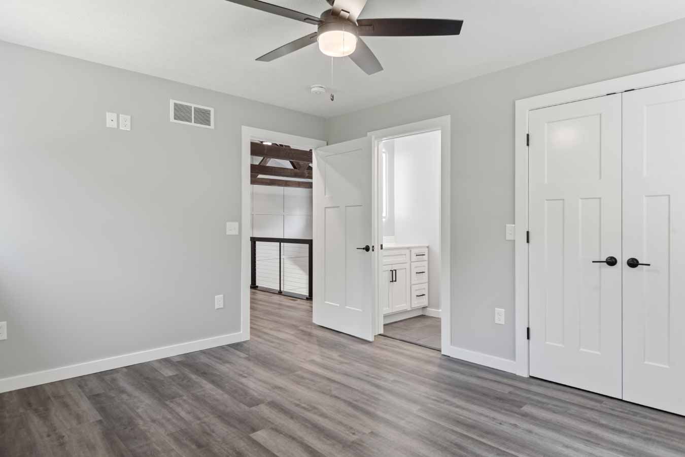 Bedroom in a modern post frame home featuring a ceiling fan, double closet doors, and a view into the shared Jack and Jill bathroom. The doorway opens to a loft with exposed wood trusses and cable railing in this stylish barndominium.