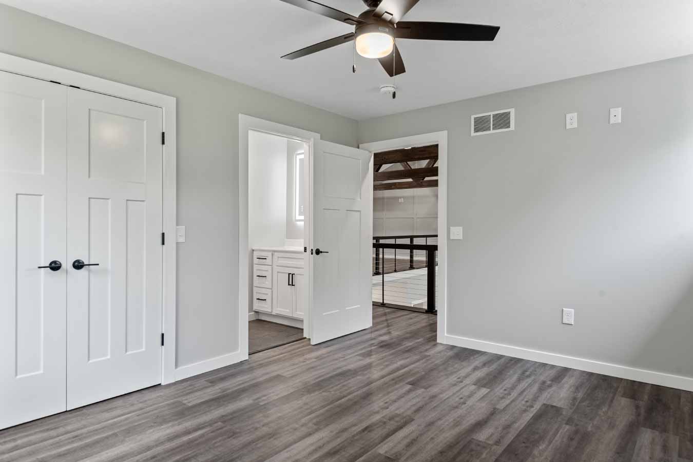 Second upstairs bedroom in a post frame house with ceiling fan, closet, and view into the Jack and Jill bathroom. The door opens to the loft with exposed trusses and cable railing.