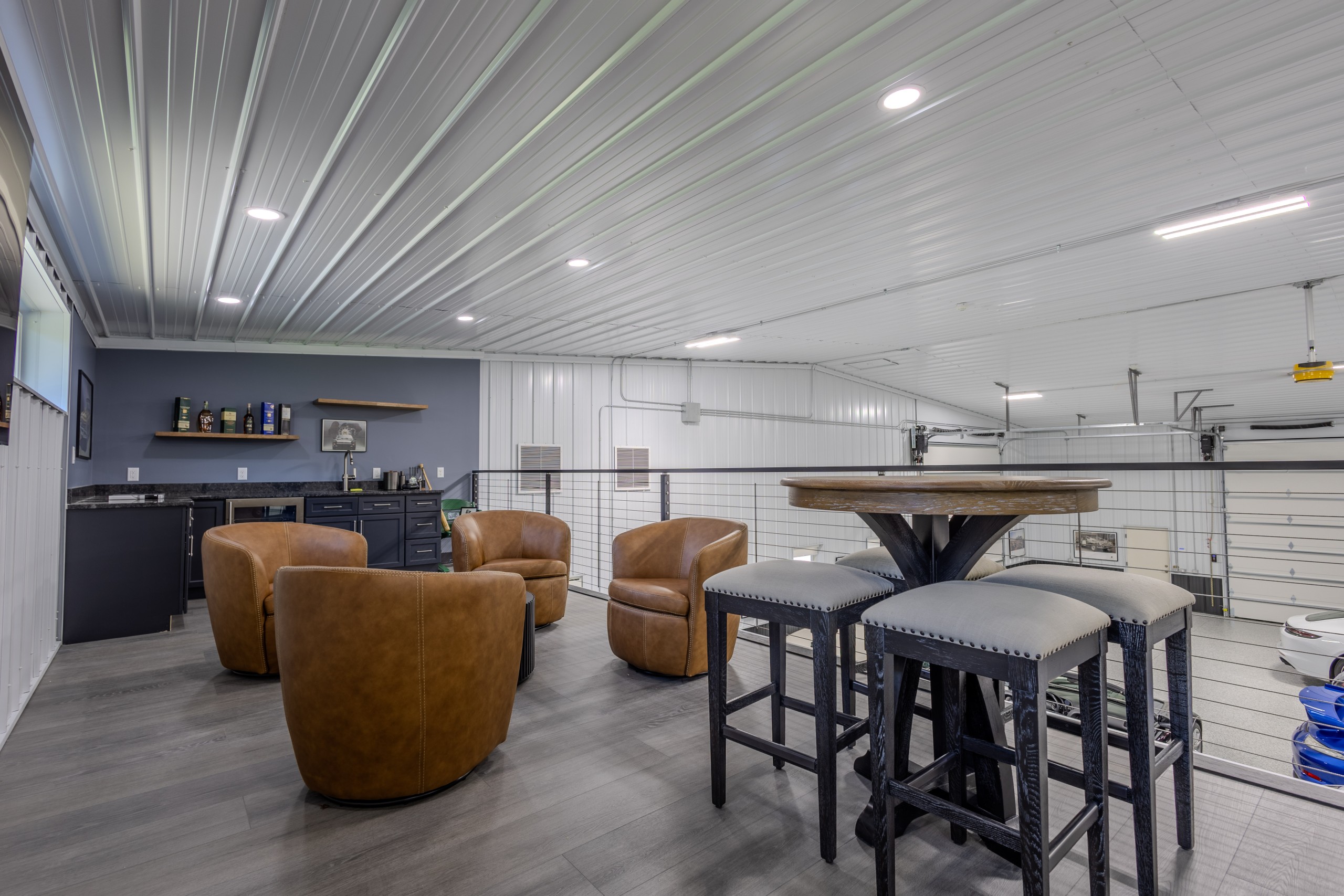 Second-floor mezzanine lounge overlooking a luxury car collection, featuring leather seating, a wet bar and modern cable railings.