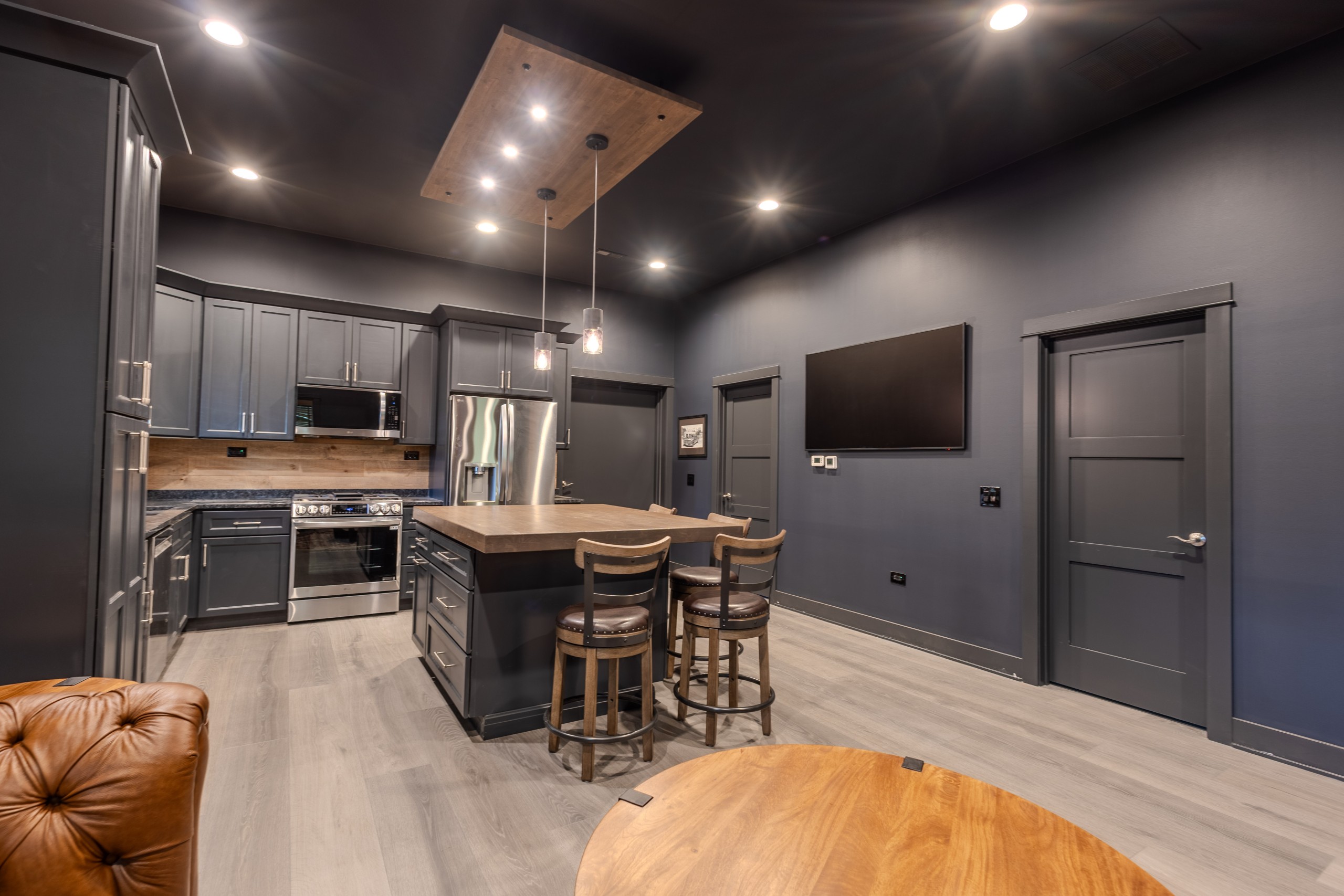 Luxury car collector garage clubhouse featuring a modern kitchen, large island with bar seating, dark cabinetry and a comfortable lounge area.